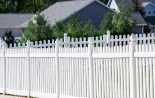 A white vinyl picket fence with decorative caps encloses a residential yard, with trees and neighbouring houses visible in the background.