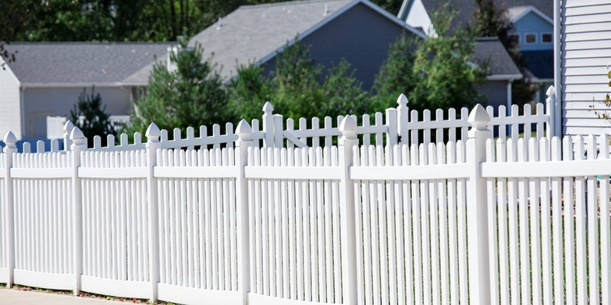 A white vinyl picket fence with decorative caps encloses a residential yard, with trees and neighbouring houses visible in the background.